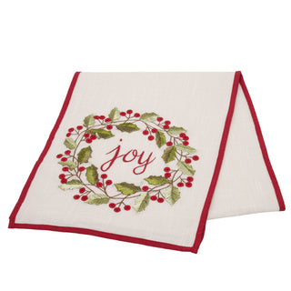An off-white linen holiday table runner with a red border and an embroidered holly wreath with the word 'joy' in red script, shown on a plain white background.