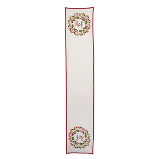 An off-white woven table runner with a thin red border, featuring two green and red holly wreath designs with the word 'Joy' in cursive script.