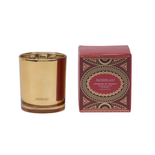 An Archipelago candle in a reflective gold glass vessel, shown next to its ornate red and gold patterned packaging box on a plain white background.