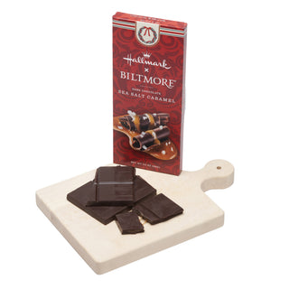 A package of Hallmark x Biltmore Sea Salt Caramel chocolate stands behind a white cutting board with several broken pieces of dark chocolate on a white background.