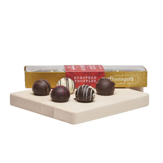 Five chocolate truffles, including dark, milk, and white chocolate varieties, displayed on a light wooden block in front of their gold and red packaging box. The brand 'Rissinger's' is visible on the box.