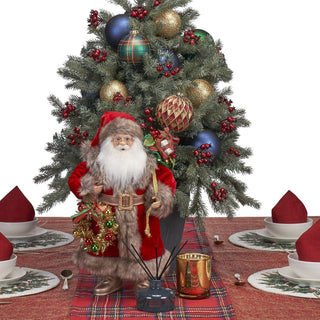 A Santa Claus figurine stands on a red plaid table runner in front of a decorated Christmas tree, with a gold candle holder, reed diffuser, and white plates with red napkins arranged around him on a white background.