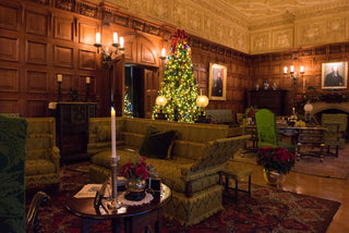 A view of Biltmore's Oak Sitting Room with dark wood paneling, ornate carved ceilings, and a decorated Christmas tree in the center. The room features vintage upholstered sofas, a patterned rug, and warm candle lighting from wall sconces.