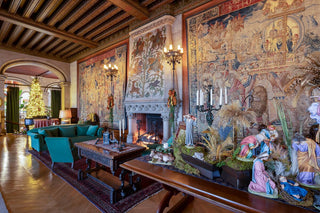 A grand, historic living room inside Biltmore House with elaborate tapestries, a fireplace, teal velvet armchairs, a dark wood coffee table, and a long wooden console table decorated with a nativity scene.