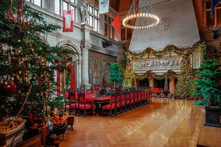 A grand, high-ceilinged interior image of Biltmore's Banquet Hall decorated for Christmas, with a large lit chandelier, polished wood floors, red upholstered chairs, and multiple decorated Christmas trees and garlands.