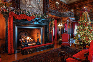 A grand, ornate view of Biltmore's Library room decorated for Christmas, showcasing a large dark fireplace burning with flames, decorated with red drapery, green garlands, and a brightly lit Christmas tree. Red upholstered chairs, dark wood paneling, and patterned rugs are visible throughout the room.
