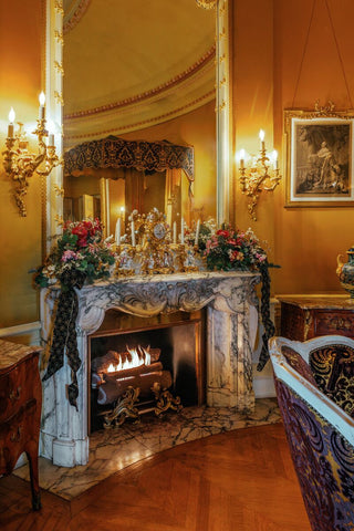 An ornate interior scene from Mrs. Vanderbilt's bedroom at Biltmore Estate, featuring a marble fireplace with a lit fire, decorated with flowers and glassware, set against warm gold walls with wall sconces and a large mirror.