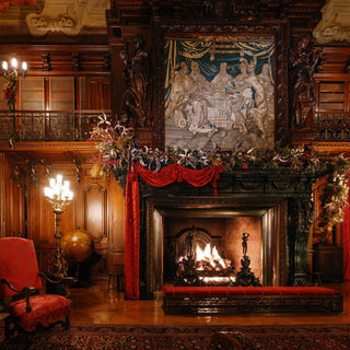 A view of Biltmore's Library room featuring a large carved fireplace, tapestry above, red drapery, a burning fire, and decorative floral garlands. A red upholstered armchair and a brass candlestick are visible in the room.