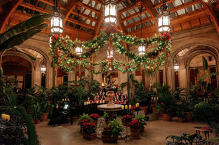 Biltmore House’s Winter Garden decorated with illuminated Christmas garlands and red bows, with a choir gathered behind the Boy with Geese sculpture.