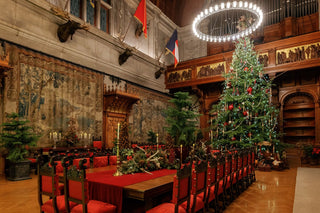 A grand interior of Biltmore House, Banquet hall is decorated with a large Christmas tree with red ornaments, surrounded by red upholstered chairs and long tables. The room features ornate wood paneling, tapestries, mounted animal heads, flags, and a large circular chandelier overhead.