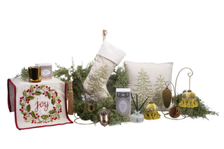 A holiday home decor collection on a white background, including a white embroidered table runner with red trim and a red 'joy' motif, a matching white Christmas stocking, a white pillow with a tree design, several gold and silver ornaments, greenery, and small decorative accessories.